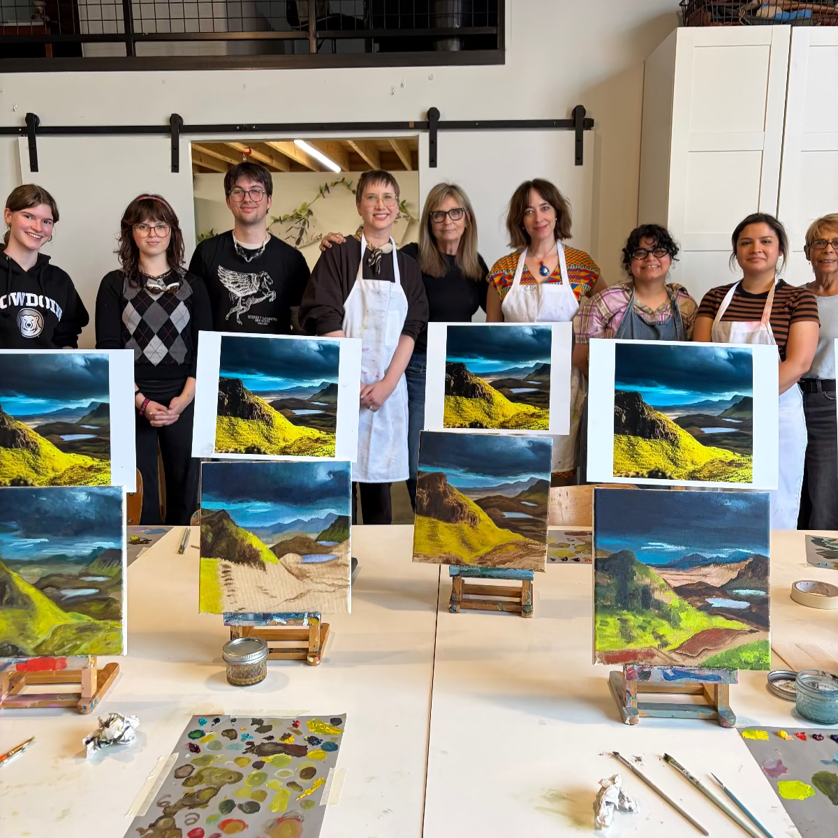 Group of people holding up their landscape paintings in a studio setting.